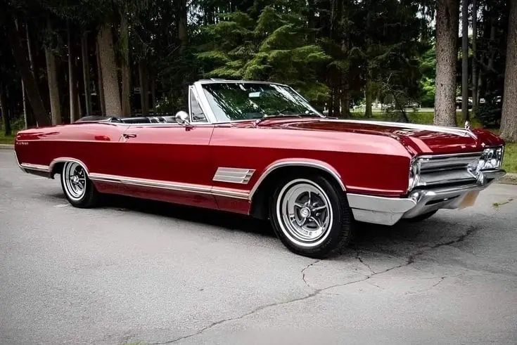 A red classic convertible car with chrome trim and whitewall tires is parked on a cracked asphalt road surrounded by tall…
