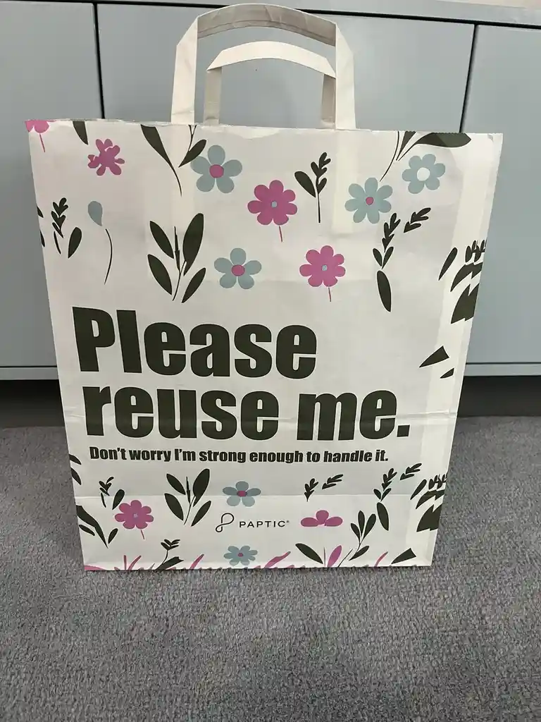 A white paper shopping bag with pink and blue floral patterns and green leaves stands upright on a gray carpet. The bag…