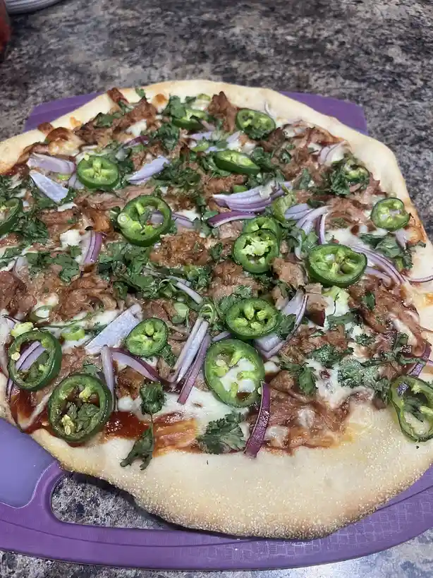 A close-up photo of a homemade pizza on a purple serving tray, topped with shredded meat, melted cheese, sliced jalapeños…