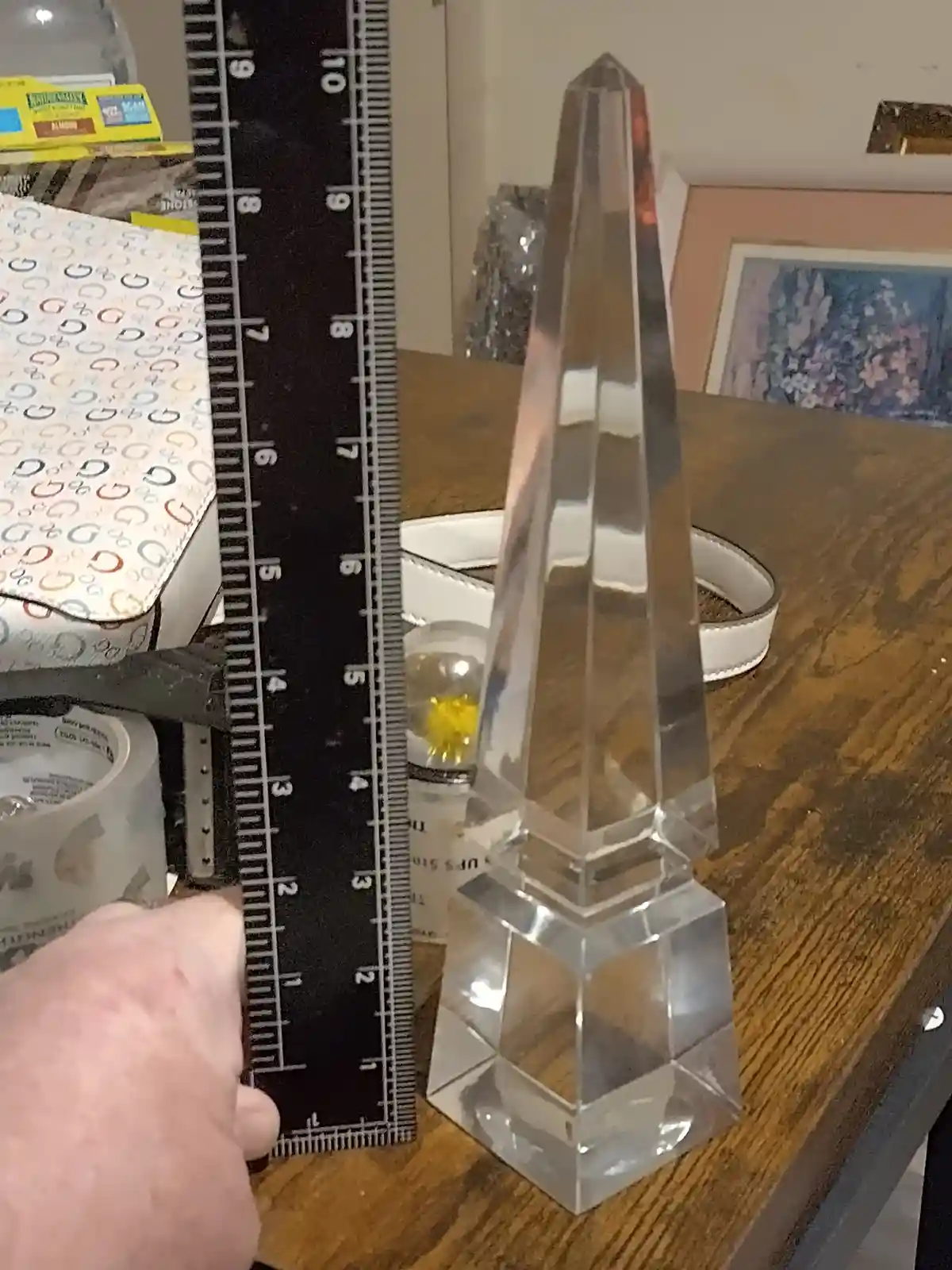 A hand holds a black ruler next to a clear, faceted crystal pyramid on a wooden table, with the ruler showing the pyramid's…