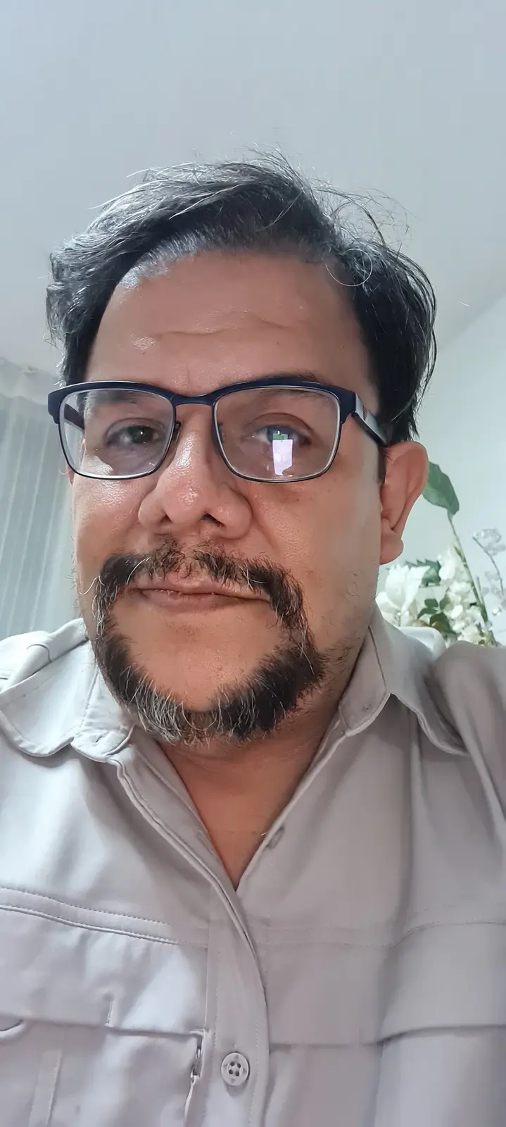 A close-up selfie of a middle-aged man with dark hair showing gray streaks, wearing black-rimmed glasses and a light-colored…