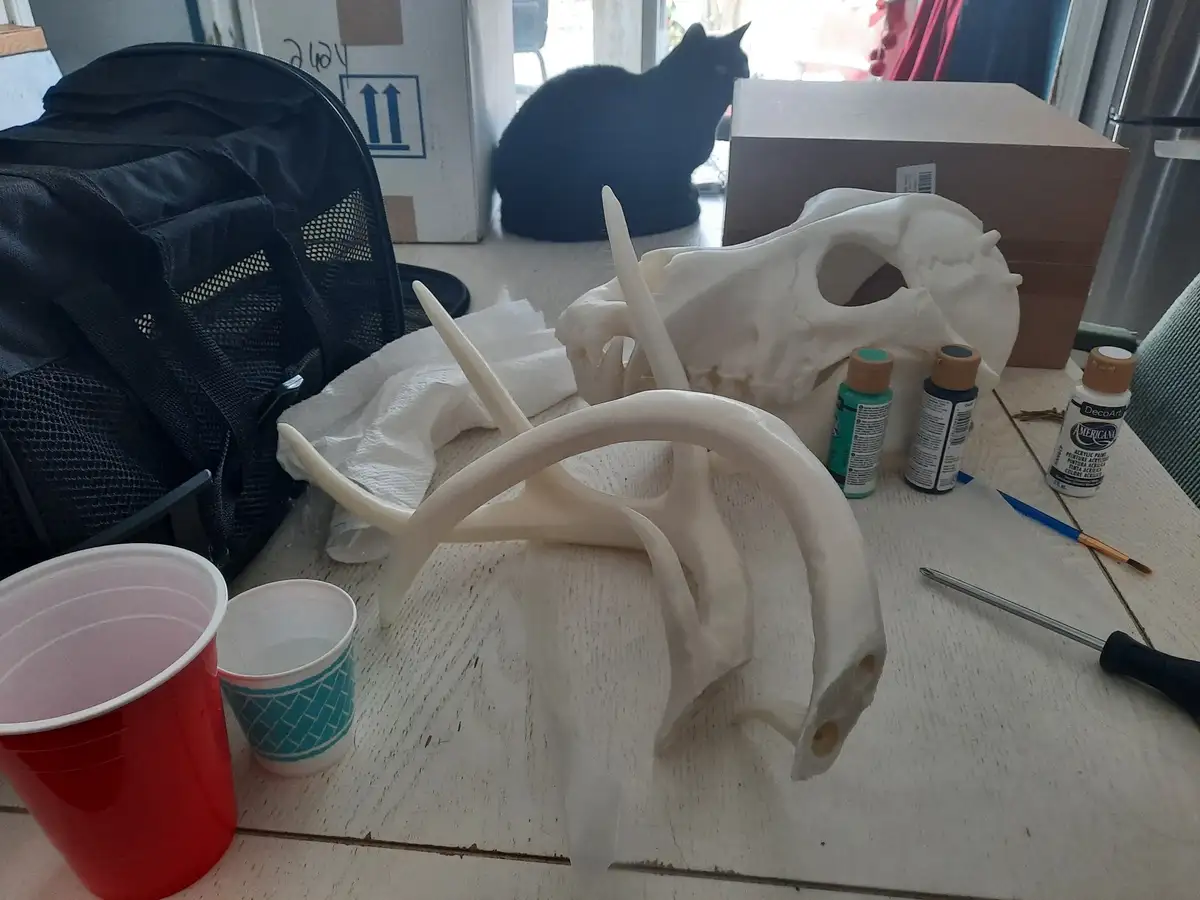 A white deer skull and antlers sit on a wooden table surrounded by craft supplies including paint bottles, brushes, and…