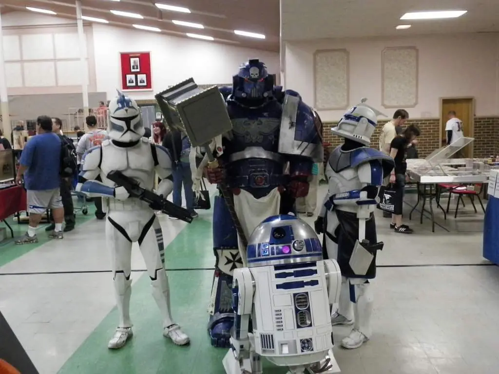 Three people in Star Wars cosplay pose together at a convention: two clone troopers flanking a towering blue Space Marine in…