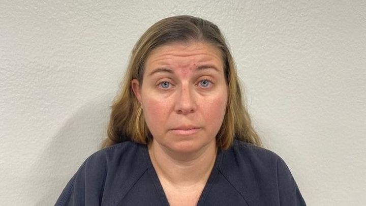 A woman with light brown hair and blue eyes, wearing dark blue scrubs, stares directly at the camera with a visibly…