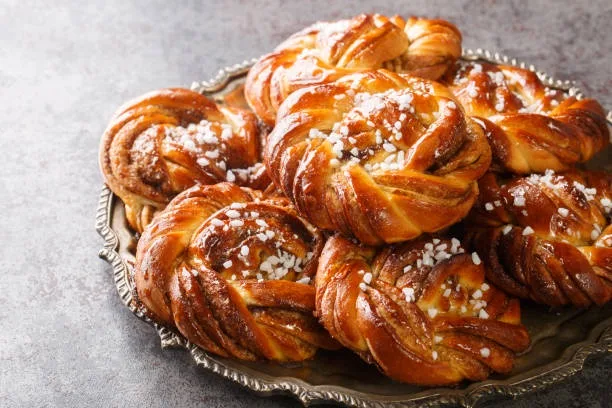 A pile of golden-brown, twisted braided pastries dusted with coarse sugar sits on an ornate silver platter against a gray…