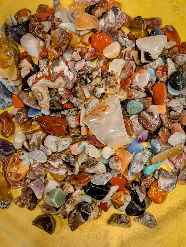 A top-down photo shows a large pile of polished, colorful stones and crystals of various shapes and sizes scattered on a…