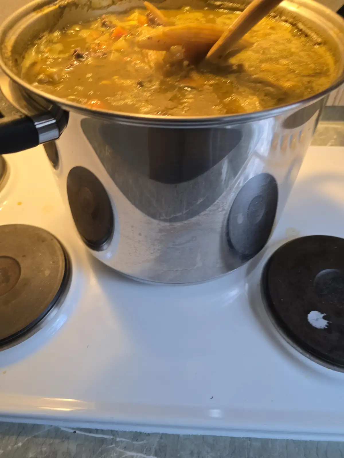 A large stainless steel pot sits on a white electric stove, filled with bubbling yellowish soup containing visible carrots…