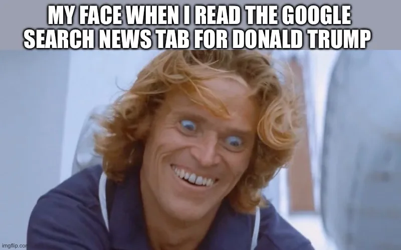 A close-up of a man with wild, curly blonde hair and an exaggerated, manic grin, his eyes wide open. He wears a dark blue…