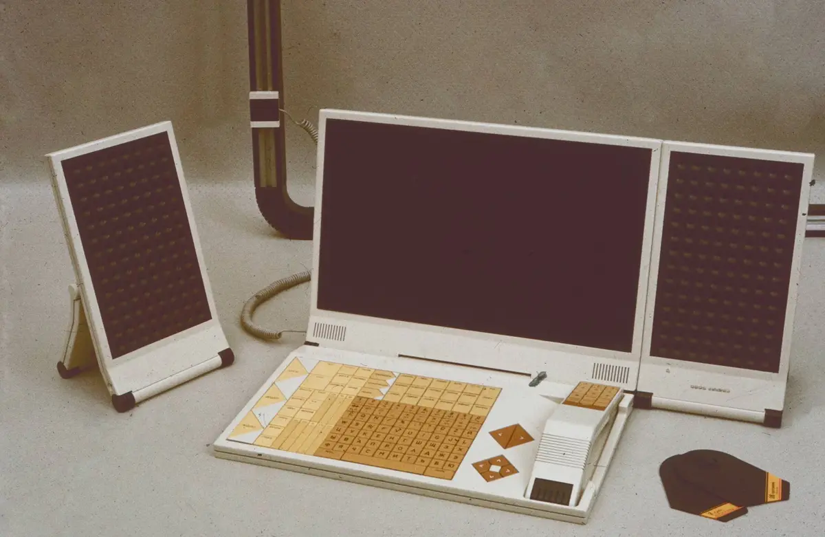 A vintage, white, retro-futuristic computer with a large central screen flanked by two small LED panels, sits on a table…