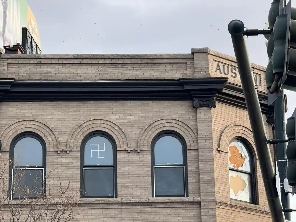 a brick building with arched windows, one of which has a white swastika drawn on it, while another window is broken with…