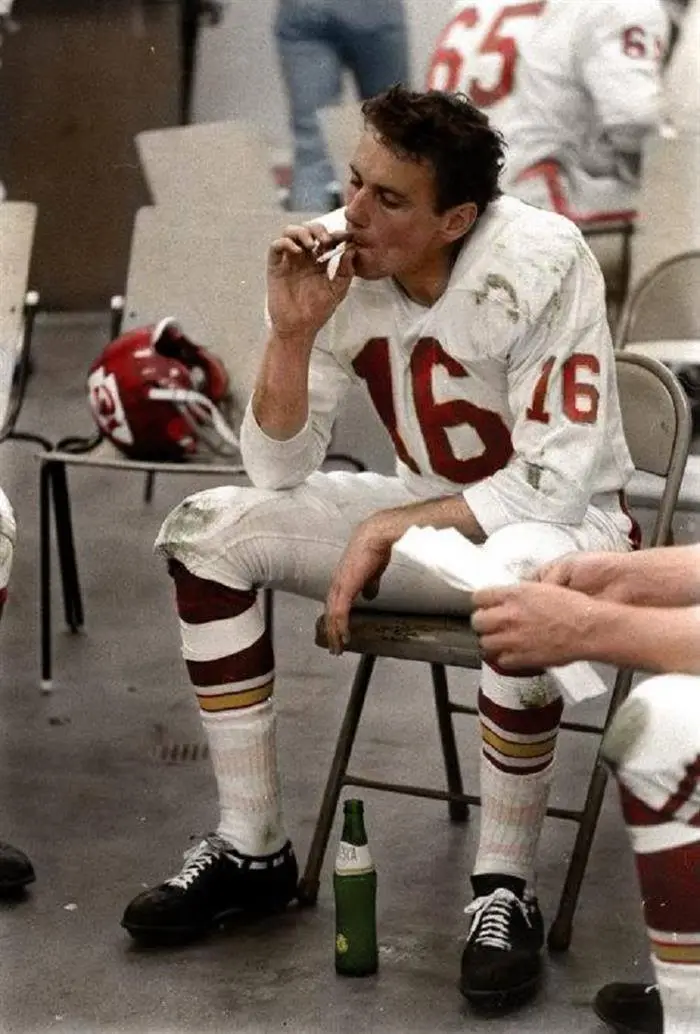 A visibly tired football player in a white jersey with red number 16 sits on a folding chair, smoking a cigarette. A green…