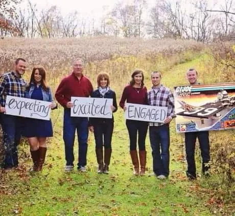 Six people stand in a grassy field holding signs that read 'Expecting', 'Excited', and 'ENGAGED', with the last person…