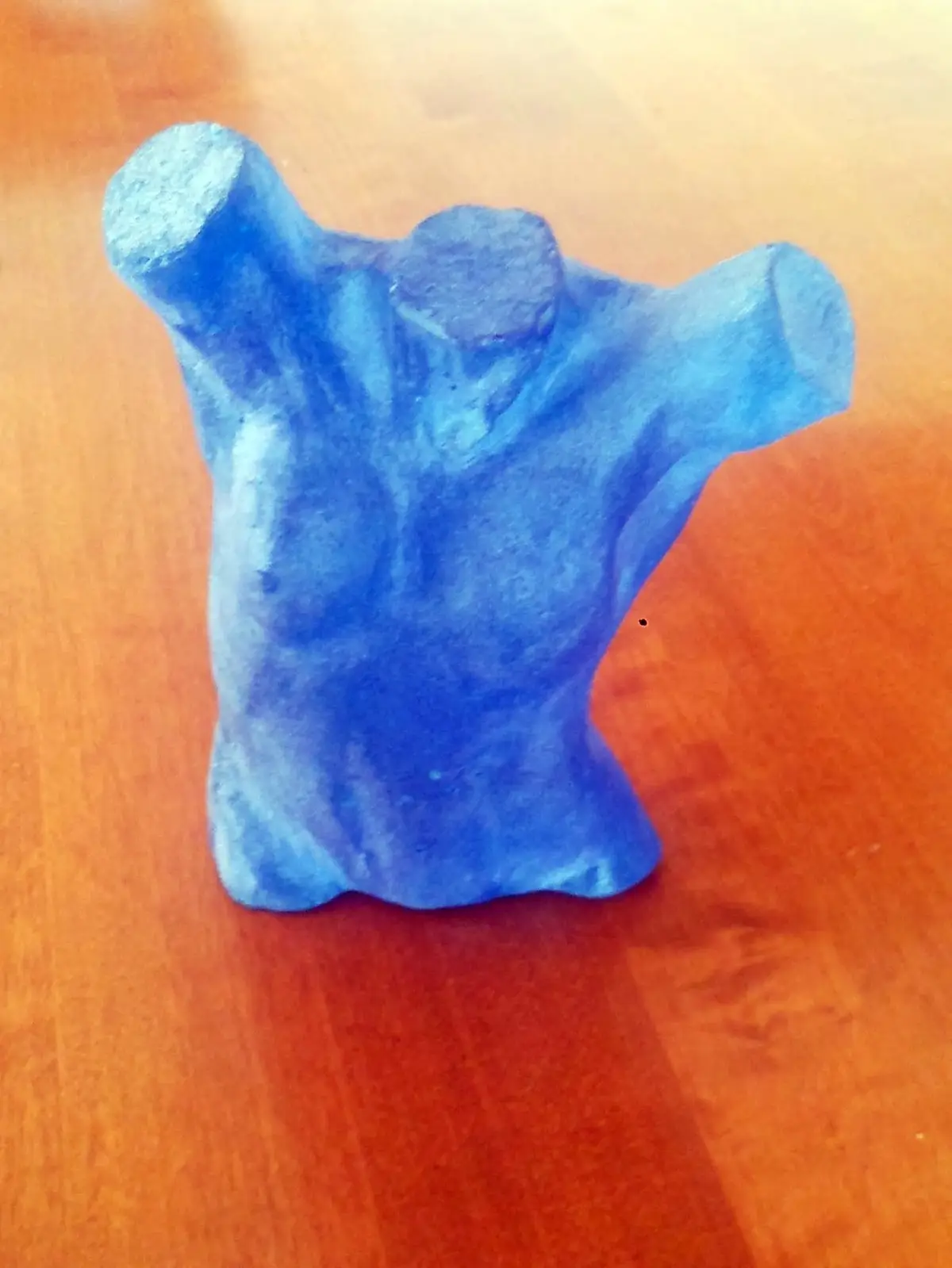 A blue, roughly sculpted object resembling a human torso with arms raised sits on a wooden surface. The overlaid text reads…