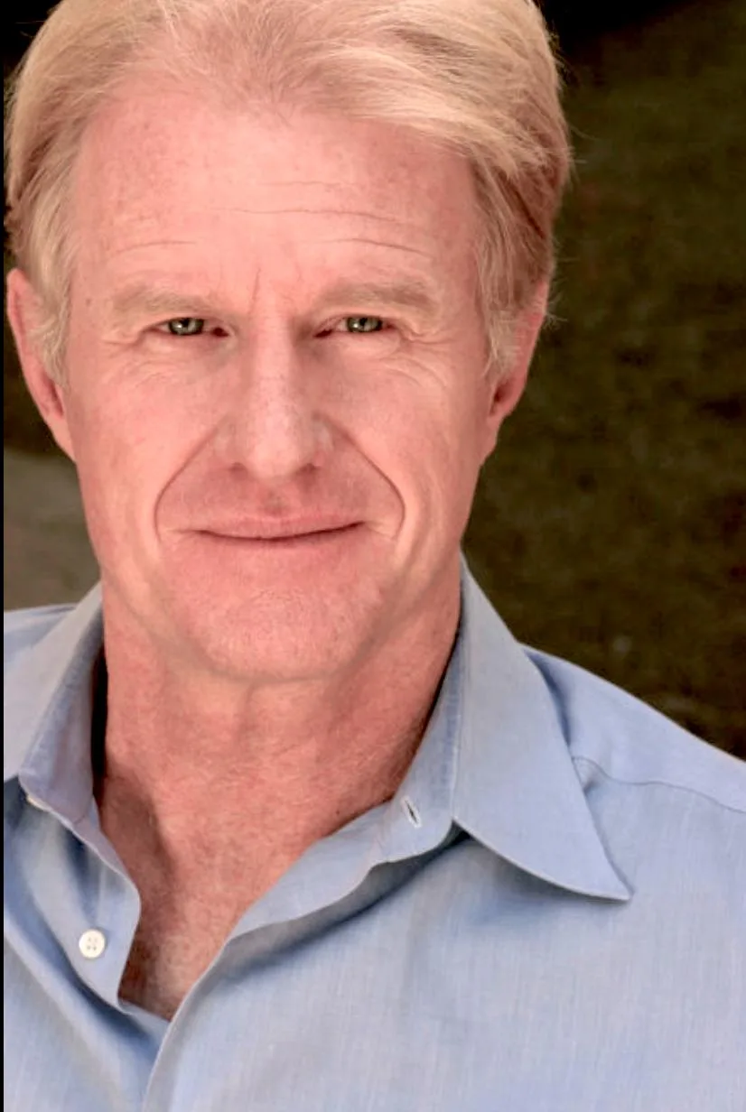 A close-up photo of a middle-aged man with blond hair and a light blue collared shirt, smiling slightly while looking…