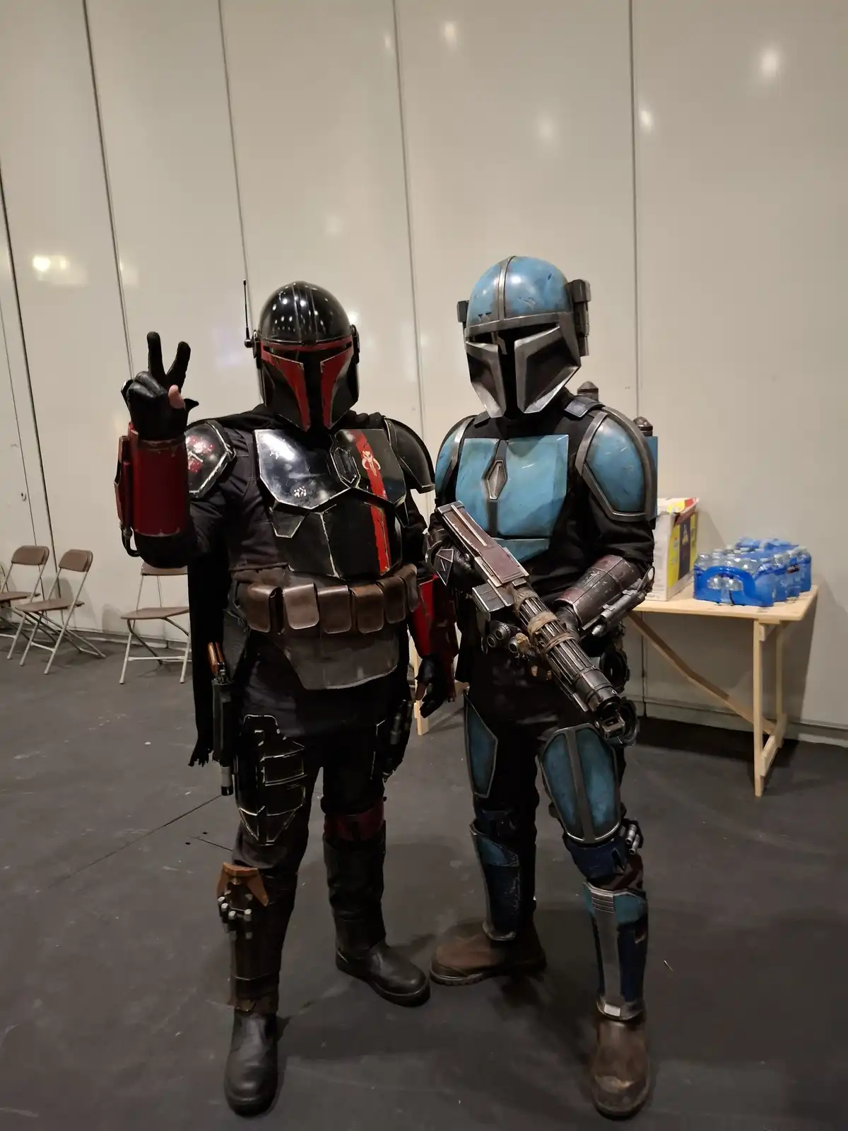 Two people in Mandalorian armor costumes stand side by side indoors; the person on the left, in black and red armor, makes a…