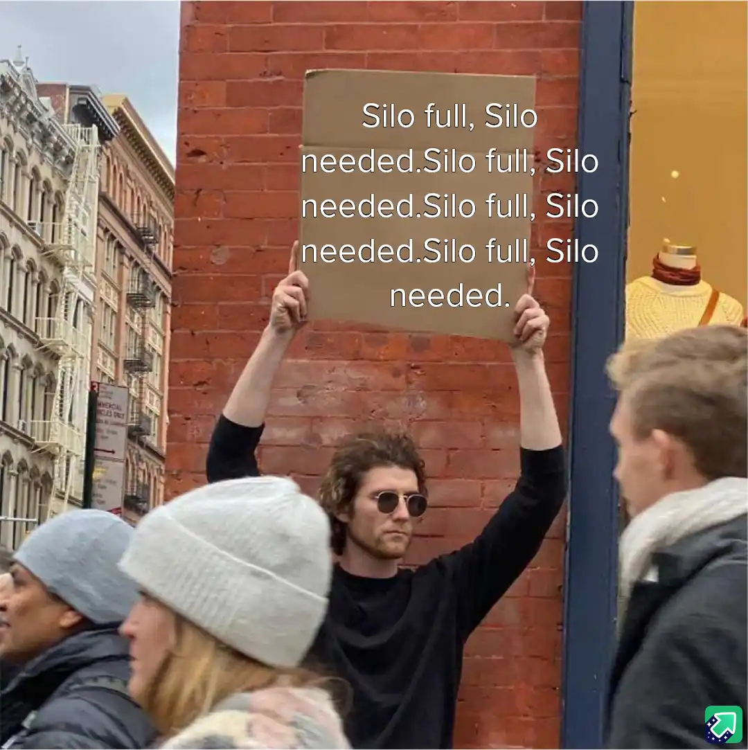 A man with curly hair and sunglasses holds up a cardboard sign repeatedly saying 'Silo full, Silo needed' while standing on…