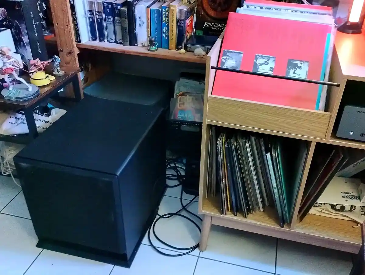 A room corner showing a large black subwoofer next to a wooden record storage unit filled with vinyl records. On a shelf…