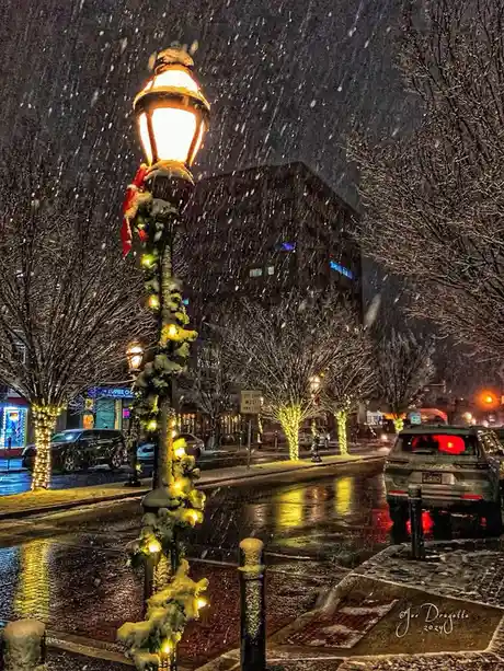 A snowy night street scene featuring a vintage-style lamppost decorated with Christmas lights, garland, and a red-and-white…