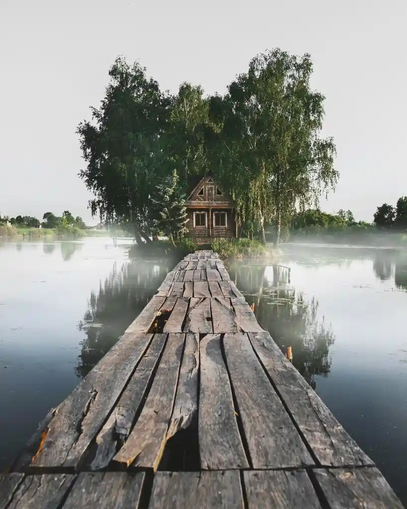 A serene, misty landscape shows a weathered wooden dock leading to a small cabin nestled among tall trees on a calm lake…