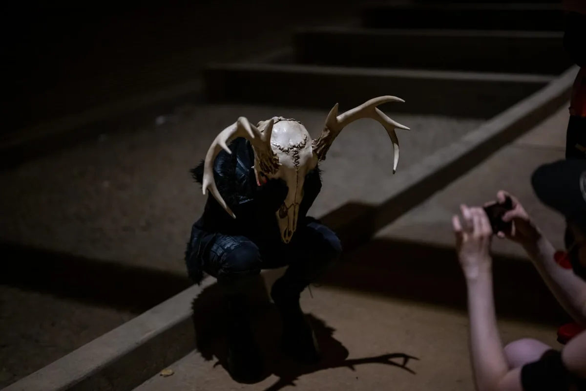 A person in a dark, dimly lit setting is crouching while wearing a large deer skull mask with antlers. Another person is…