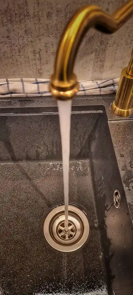 A close-up photo shows water flowing from a gold-colored faucet into a dark sink drain. The faucet has a curved spout and is…