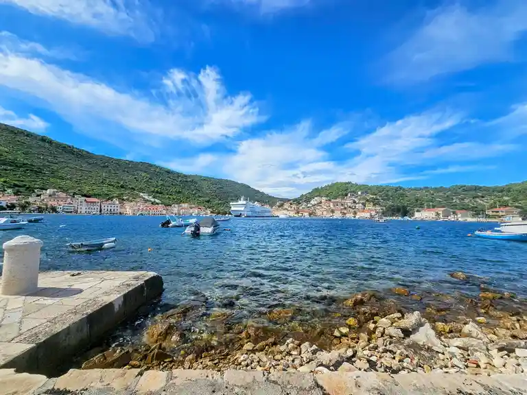 A scenic coastal view shows a harbor with boats, a large ferry named 'JADROLINIJA', and a town on a hillside under a bright…