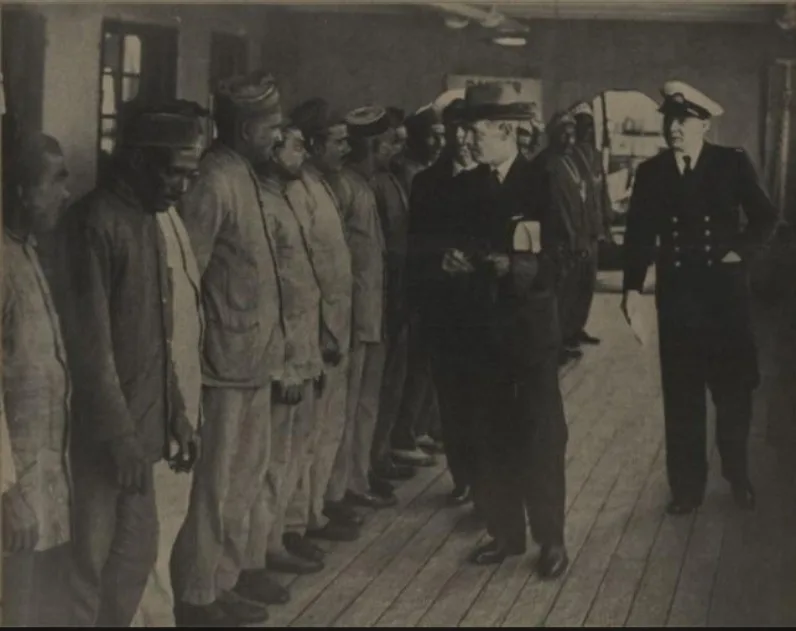 A black-and-white historical photograph shows a line of men in uniform standing on a wooden deck, facing a man in a…
