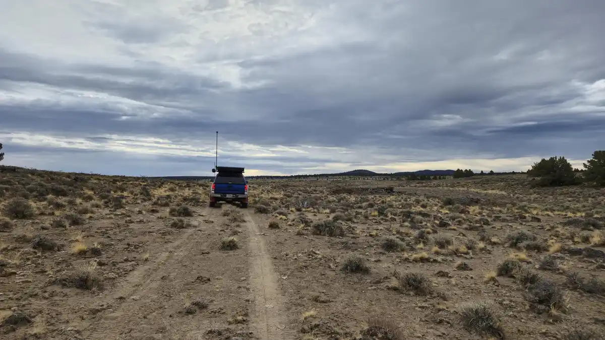 A blue pickup truck with a roof rack and antenna drives down a dirt road through a vast, arid landscape under a cloudy sky…
