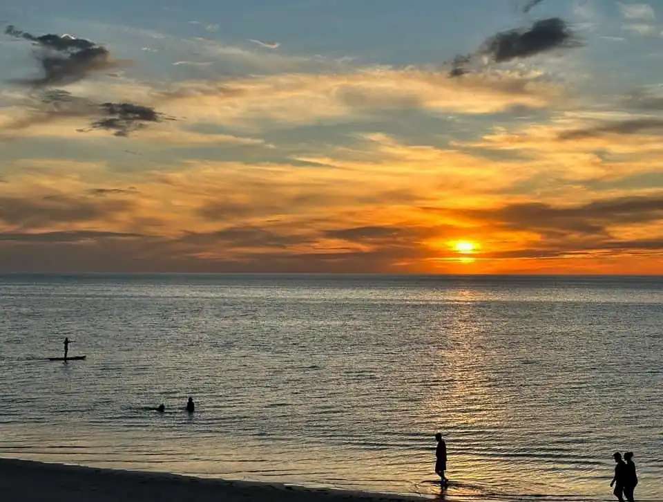A serene sunset over the ocean with silhouettes of people enjoying the beach: one person paddleboarding, two swimming, and…