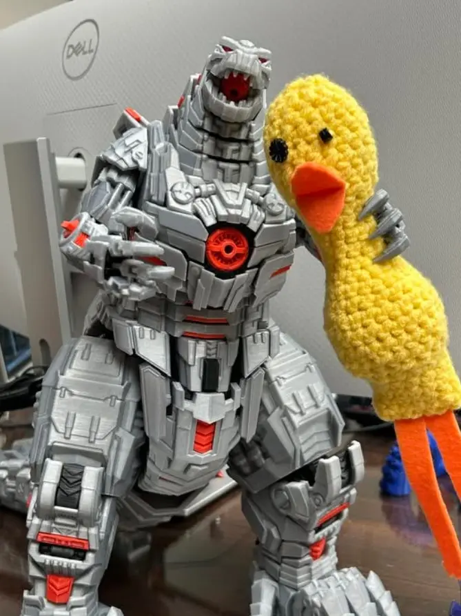 A large, grey and red mechanical Godzilla action figure stands with its mouth open, holding a bright yellow crocheted duck…