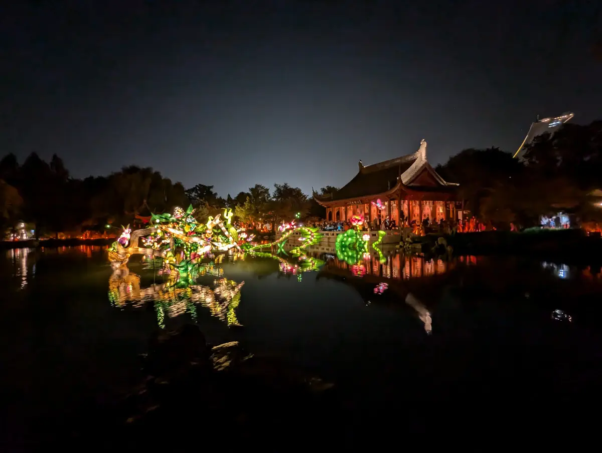 A nighttime photo of a traditional Chinese pavilion beside a pond, illuminated by colorful lanterns shaped like dragons and…