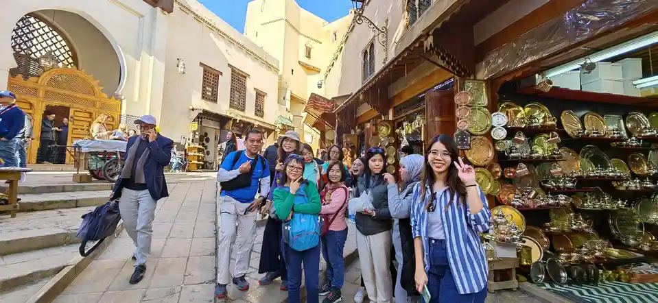 A group of smiling tourists poses for a photo in a bustling, narrow market street with ornate architecture and a shop…