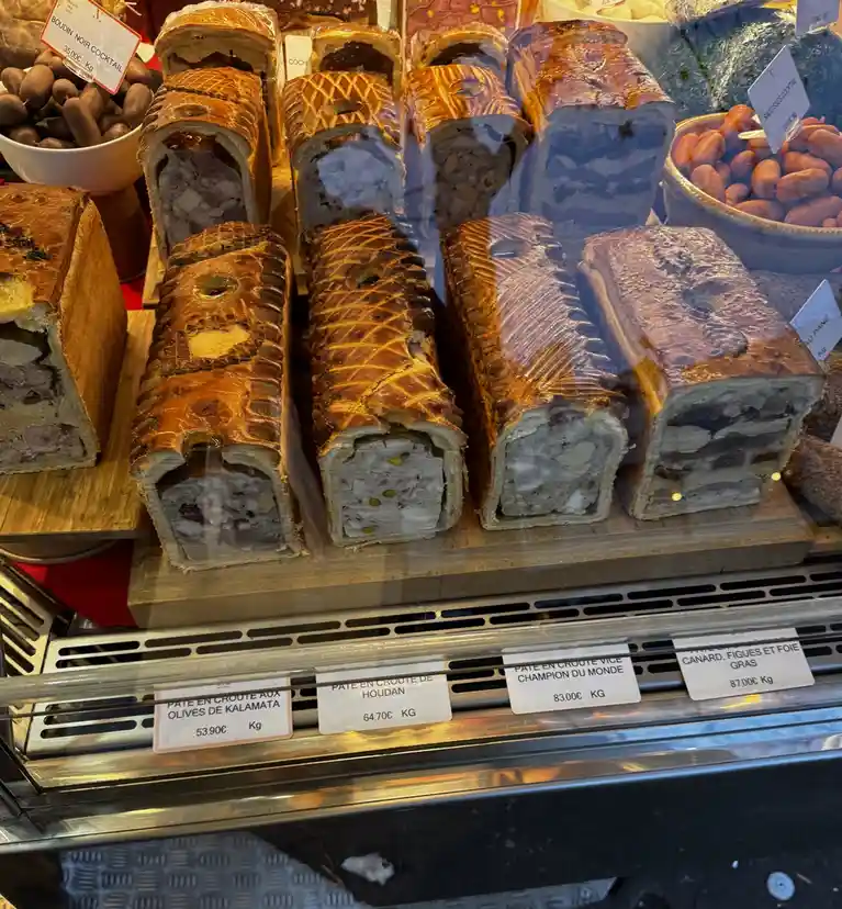 A display case shows rows of sliced pâté en croûte pastries with golden-brown crusts and visible fillings, priced per…