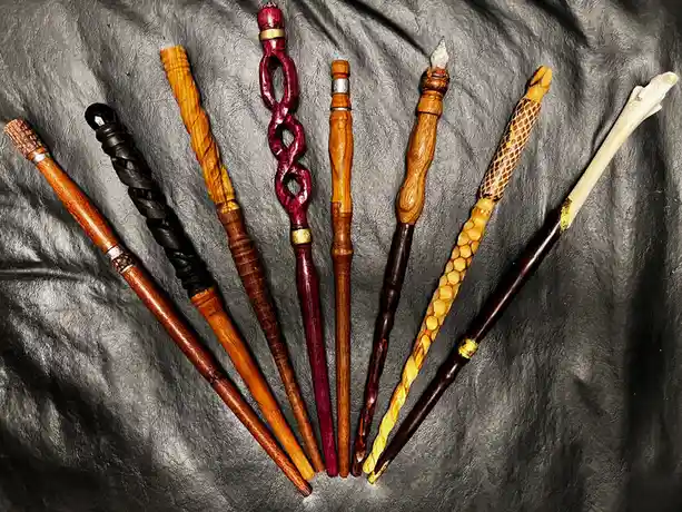 An overhead shot displays eight intricately crafted wooden wands with varied designs, colors, and embellishments, arranged…