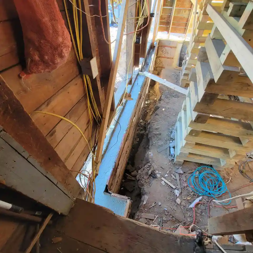 A high-angle photo of a messy, unfinished construction site showing exposed wooden framing, a trench dug into the dirt…