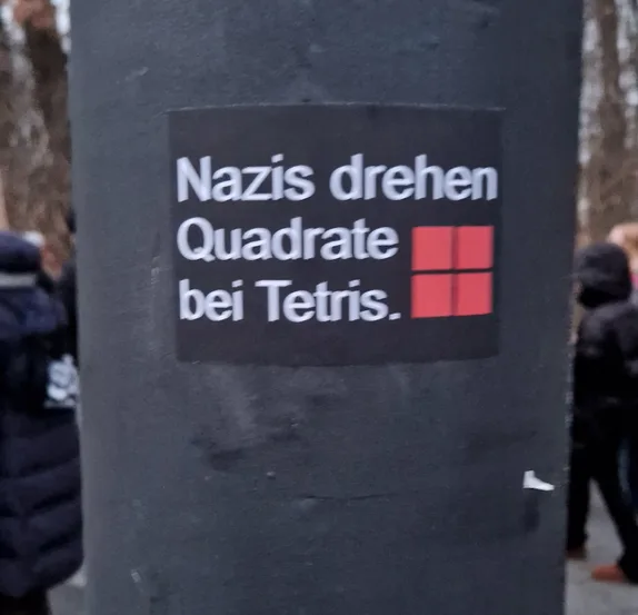 A black sticker on a dark pole shows white text reading 'Nazis drehen Quadrate bei Tetris.' alongside a red Tetris block…