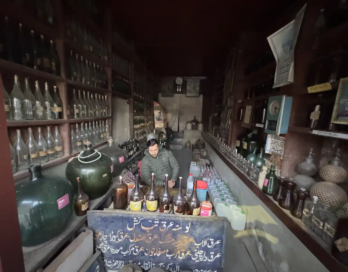 A man sits behind a counter in a dimly lit, narrow shop filled with shelves of glass bottles and large green jars. The…