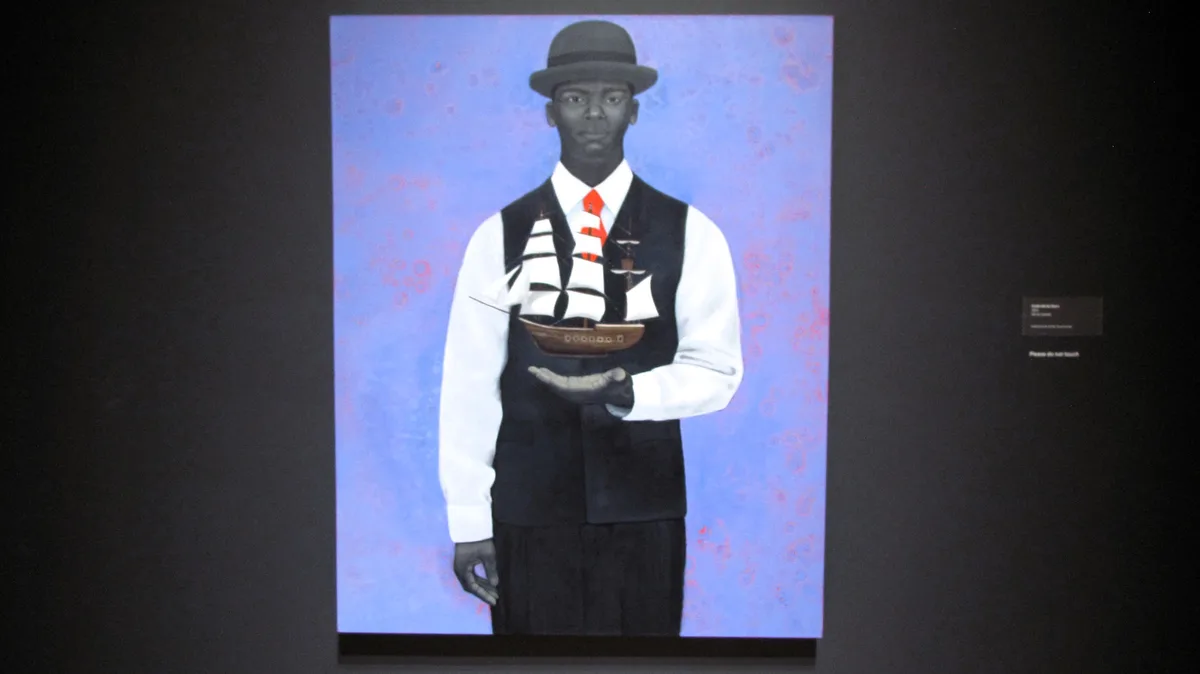 A man in a bowler hat, white shirt, red tie, and black vest holds a model ship in his cupped hands against a blue background…