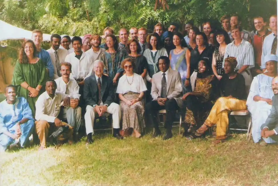 A large group of diverse people posing for a photo outdoors on grass, with some seated in front and others standing behind…