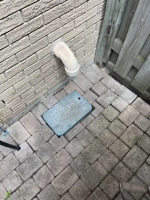 An outdoor photo shows a gray plastic drain pipe protruding from a beige brick wall, with a gray metal access cover set into…