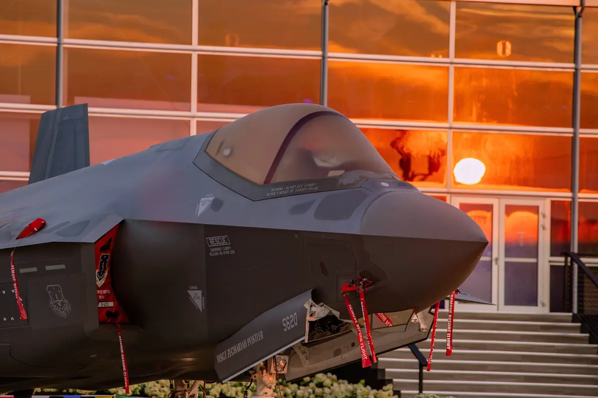 A gray F-35 fighter jet is displayed outdoors in front of a modern glass building during sunset, with red 'REMOVE BEFORE…