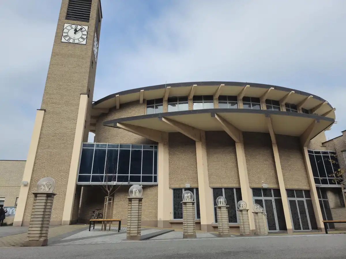 An exterior photo of a modern brick church with a tall clock tower showing 3:00 pm. The building has large windows, a curved…