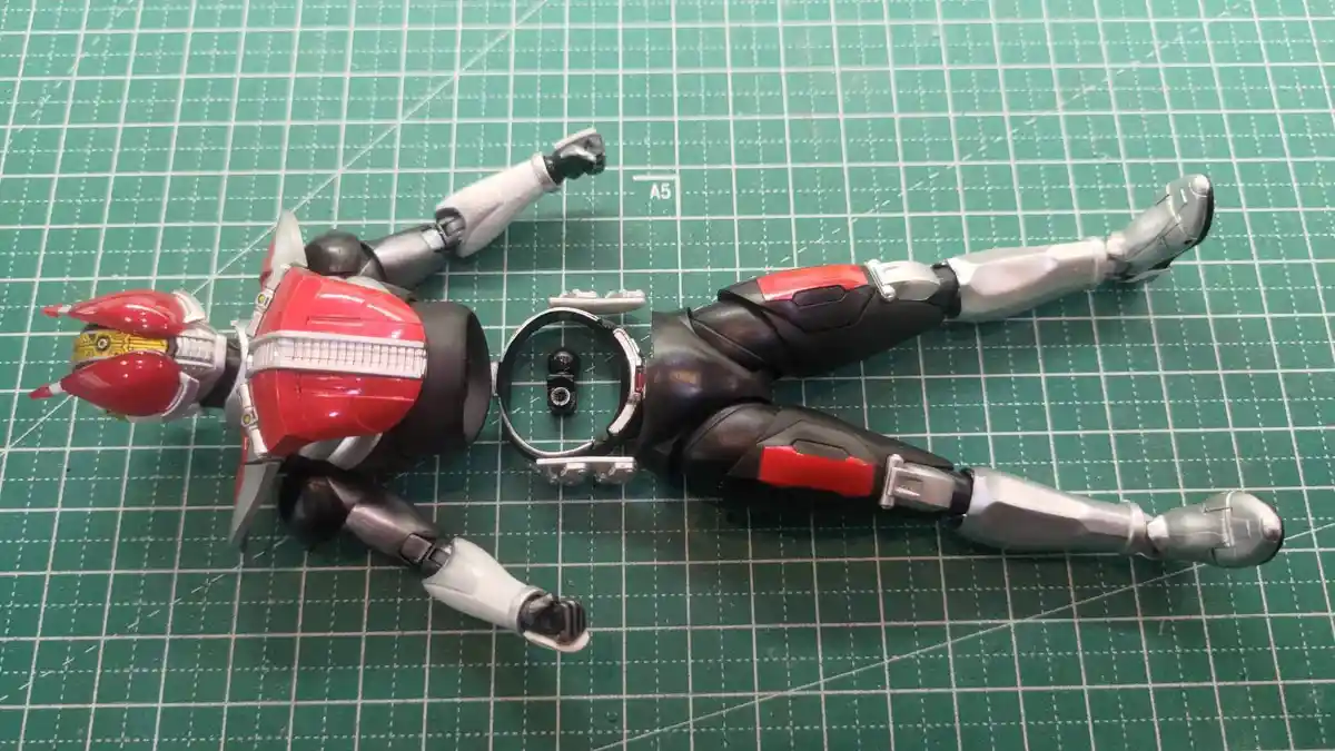 A red and silver Kamen Rider action figure lies on its back on a green cutting mat, with its torso separated from its lower…