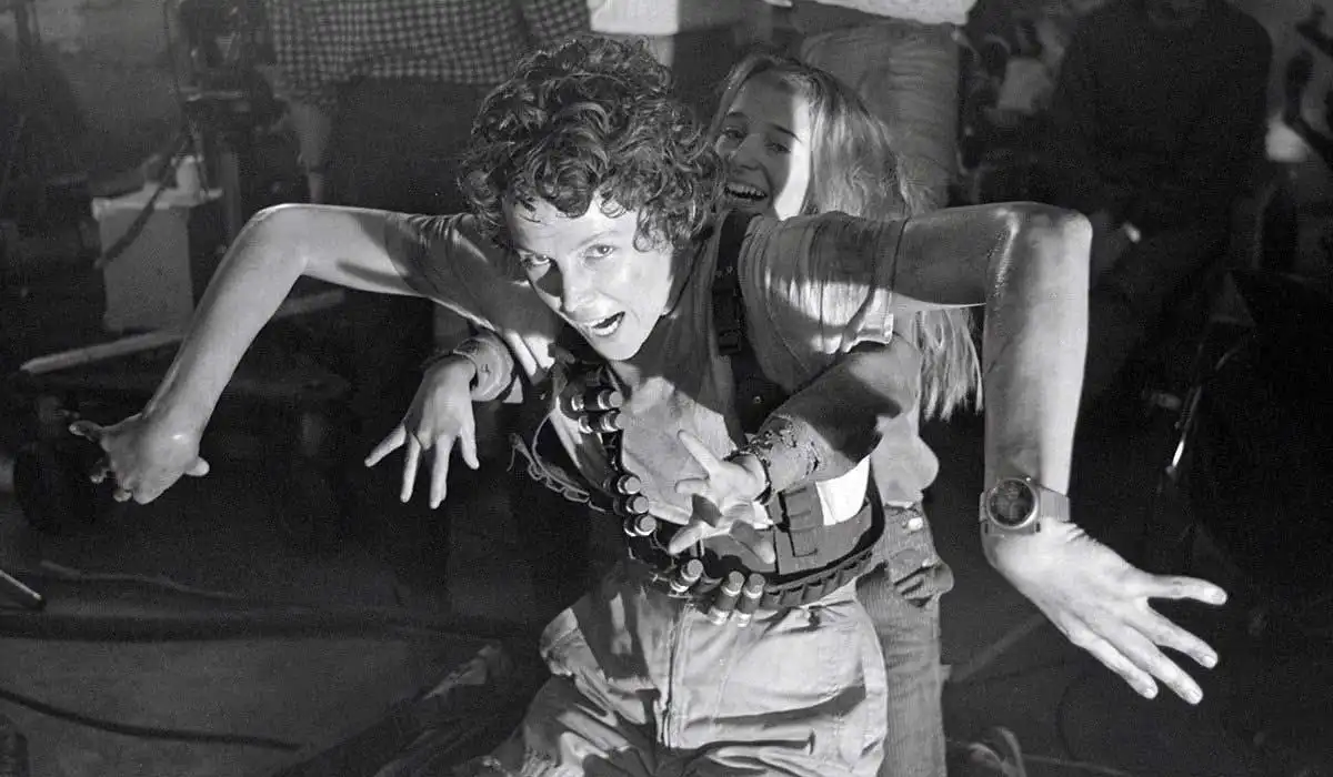 A black-and-white photo shows actress Sigourney Weaver in a tense, crouched pose on a movie set, wearing a utility belt with…