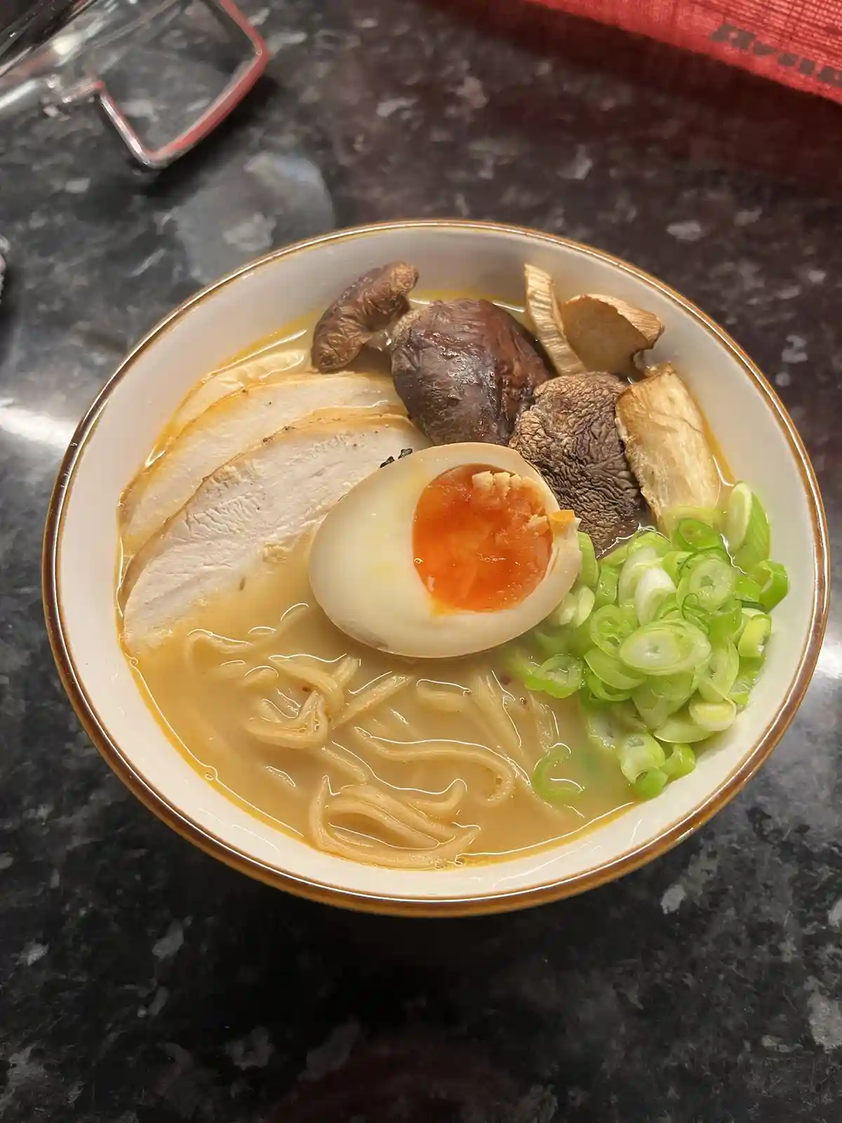 a top-down photo of a bowl of ramen on a dark countertop, featuring noodles in broth, a halved soft-boiled egg with a runny…