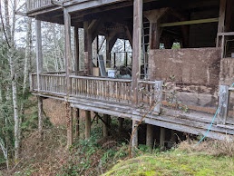 A dilapidated wooden porch of a house on stilts, surrounded by overgrown vegetation and trees, with visible decay and…
