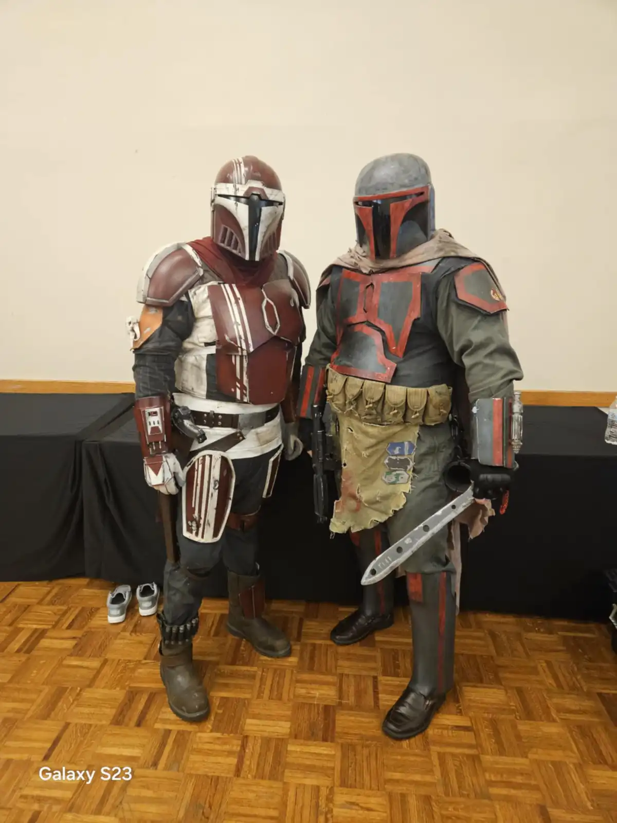 Two people in detailed Mandalorian armor costumes stand side-by-side indoors on a wooden floor, one in red and white armor…