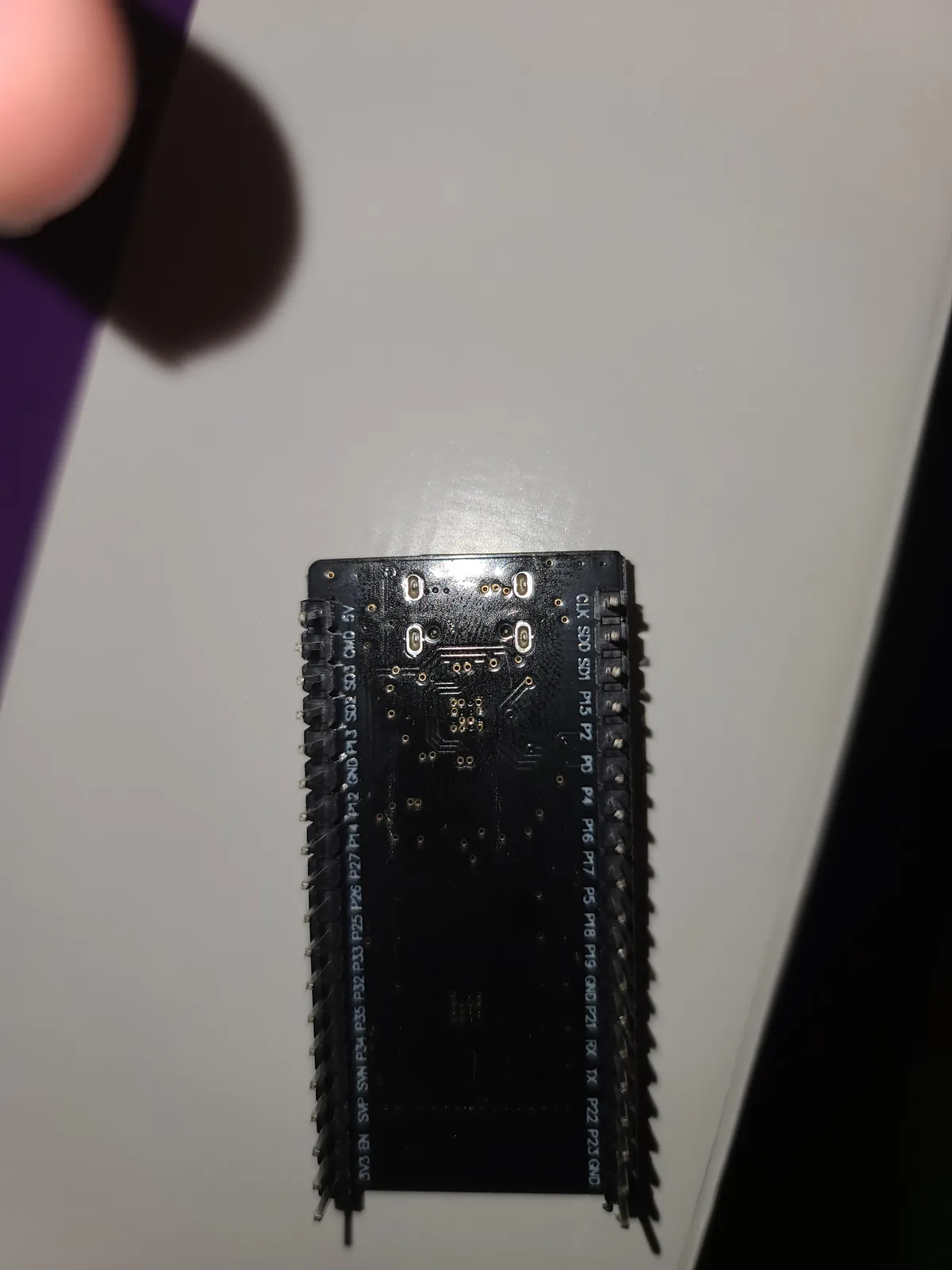 A close-up photo shows the back of a small black electronic circuit board with labeled pins along its edges, resting on a…