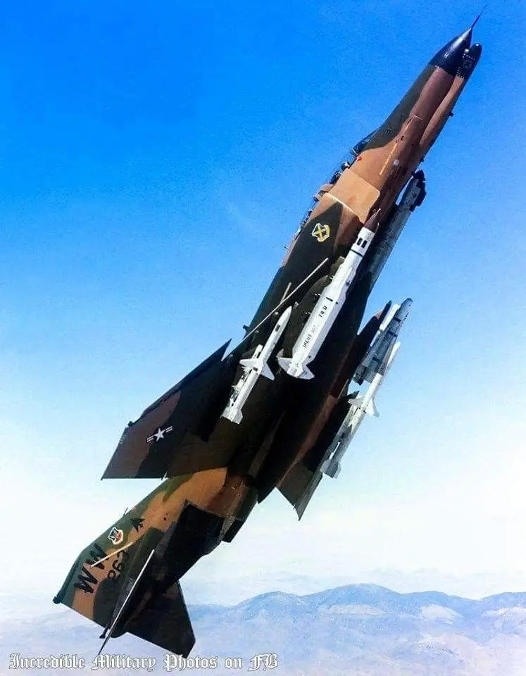 A military fighter jet, painted in camouflage, is shown flying steeply upward against a clear blue sky with distant…