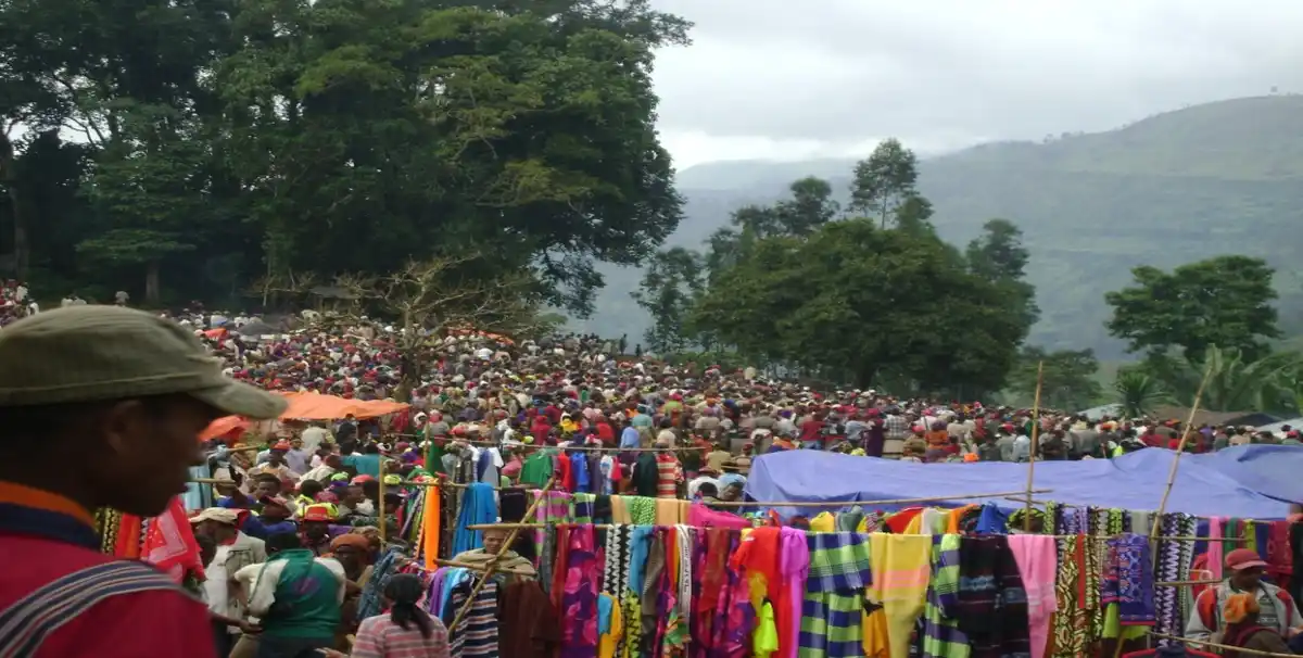 A crowded outdoor market in a lush, hilly region with many people browsing colorful textiles hanging on racks. In the…